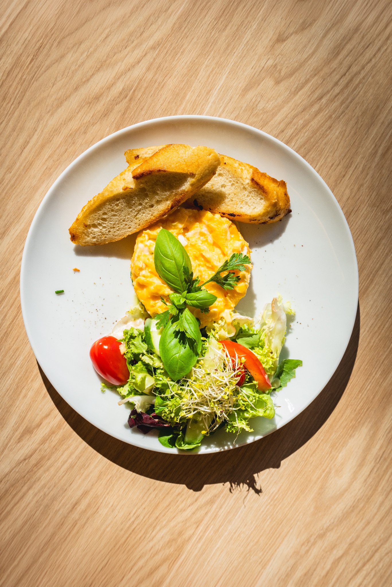 Elegancko podane śniadanie: jajecznica uformowana w kopiec z bazylią, sałatka ze świeżych warzyw i grzanki na białym talerzu na drewnianym stole w słoneczny dzień.