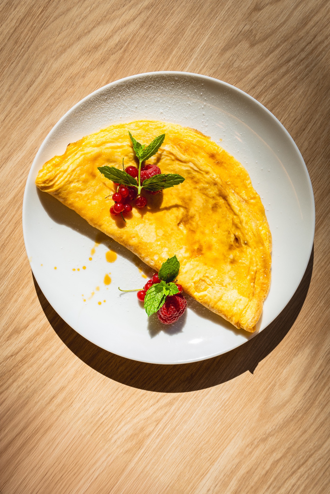 Omelet na białym talerzu posypany cukrem pudrem, udekorowany malinami, porzeczkami i listkami mięty, leżący na drewnianym stole w jasnym świetle.
