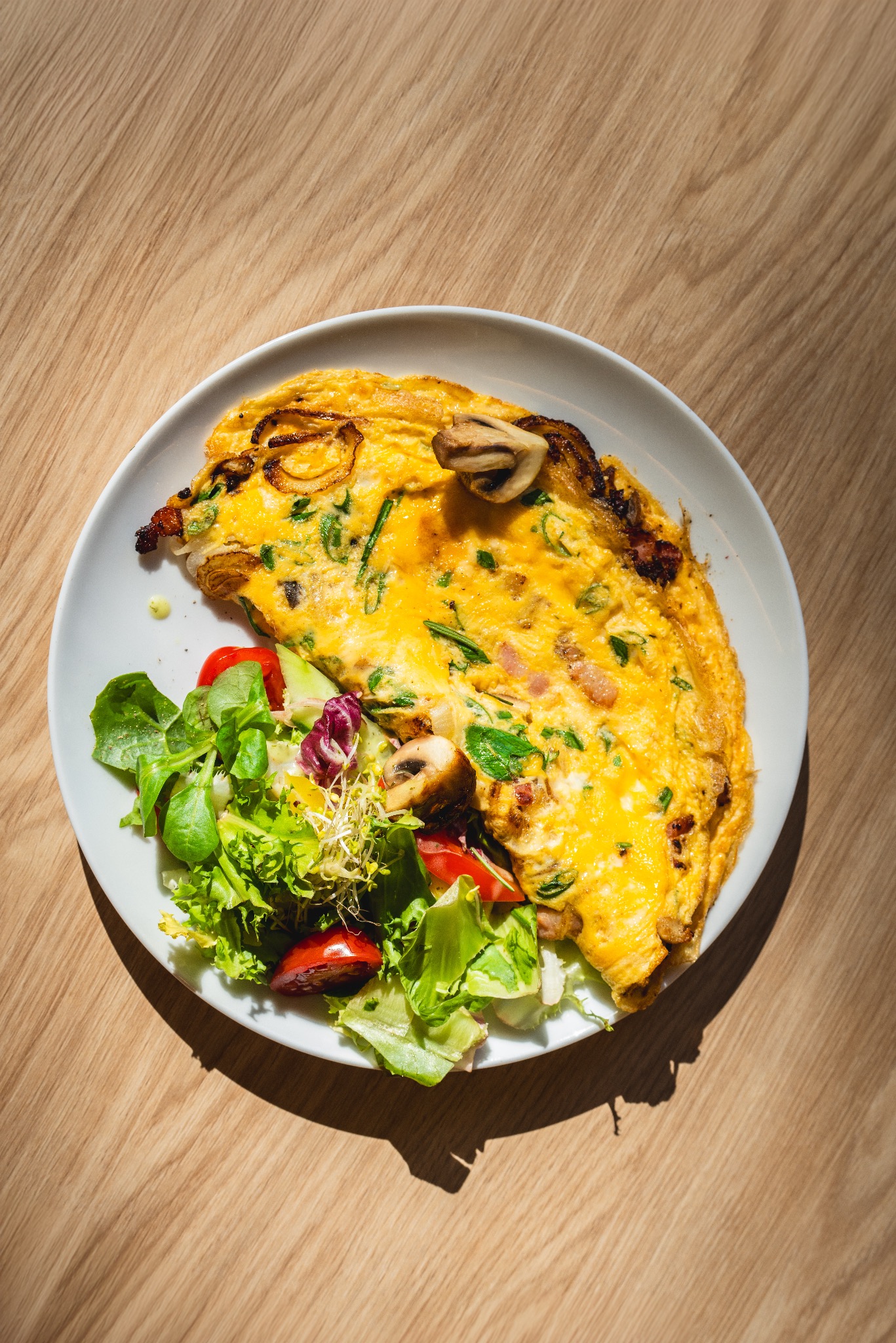 Omlet z grzybami, cebulą i ziołami, podany z sałatką ze świeżych warzyw na białym talerzu, na drewnianym stole w jasnym świetle słonecznym.