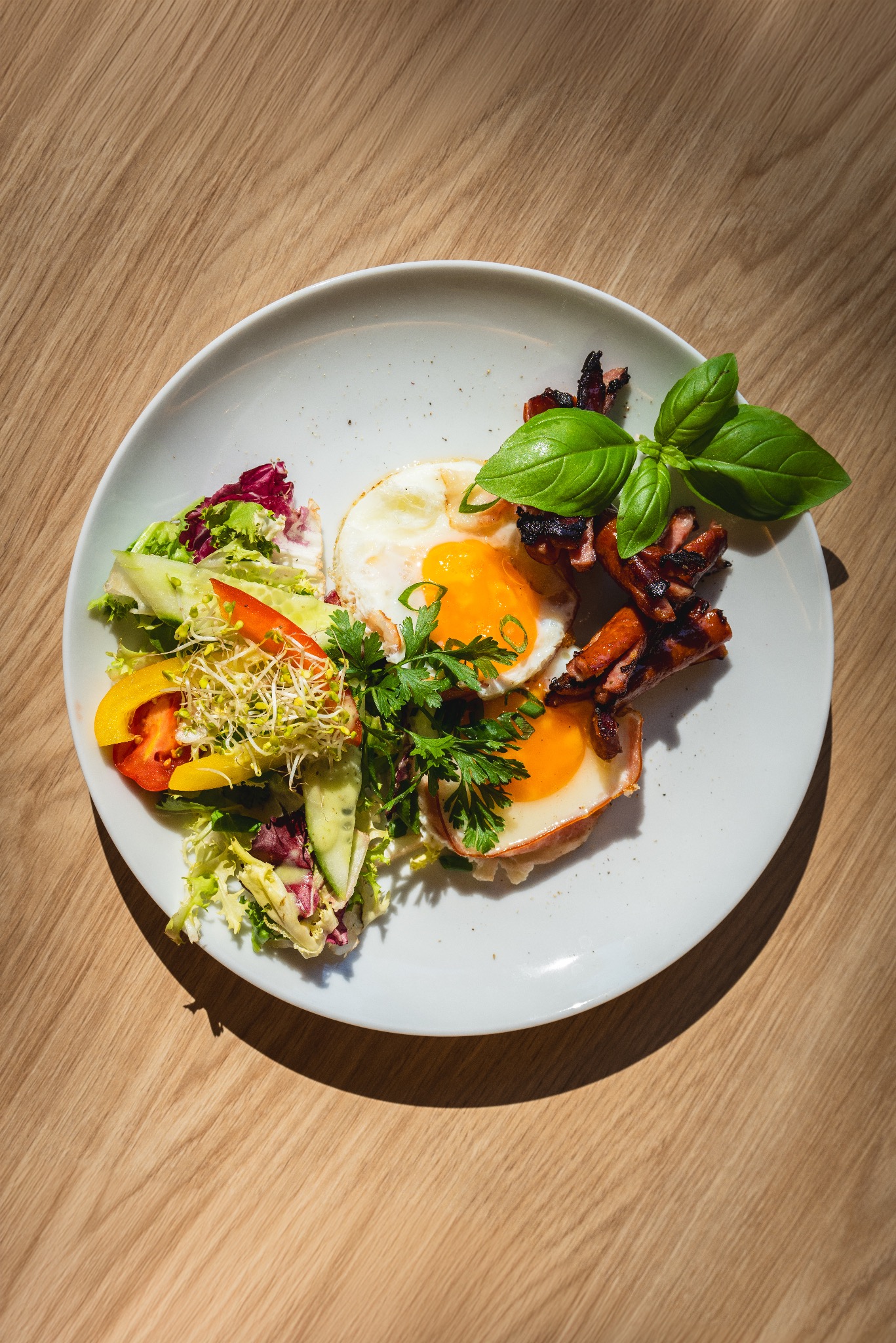 Talerz z jajkami sadzonymi, sałatką warzywną z kiełkami i smażoną kiełbasą, udekorowany bazylią i pietruszką, na drewnianym stole w naturalnym świetle.