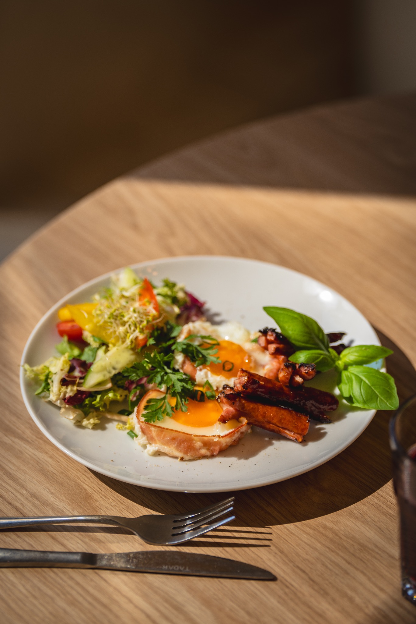 Apetyczna kompozycja śniadaniowa na białym talerzu: jajka sadzone otulone szynką, grillowane kiełbaski, świeża sałatka warzywna z kiełkami oraz gałązka bazylii, ułożone na drewnianym stole obok...