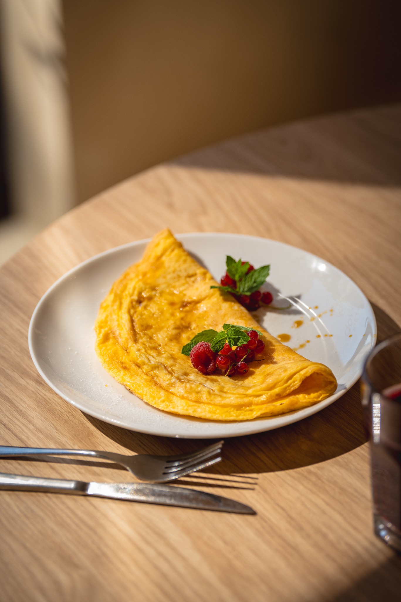 Delikatny omlet na białym talerzu, udekorowany świeżą miętą i czerwoną porzeczką, polany syropem, podany na jasnym drewnianym stole z widelcem i nożem.