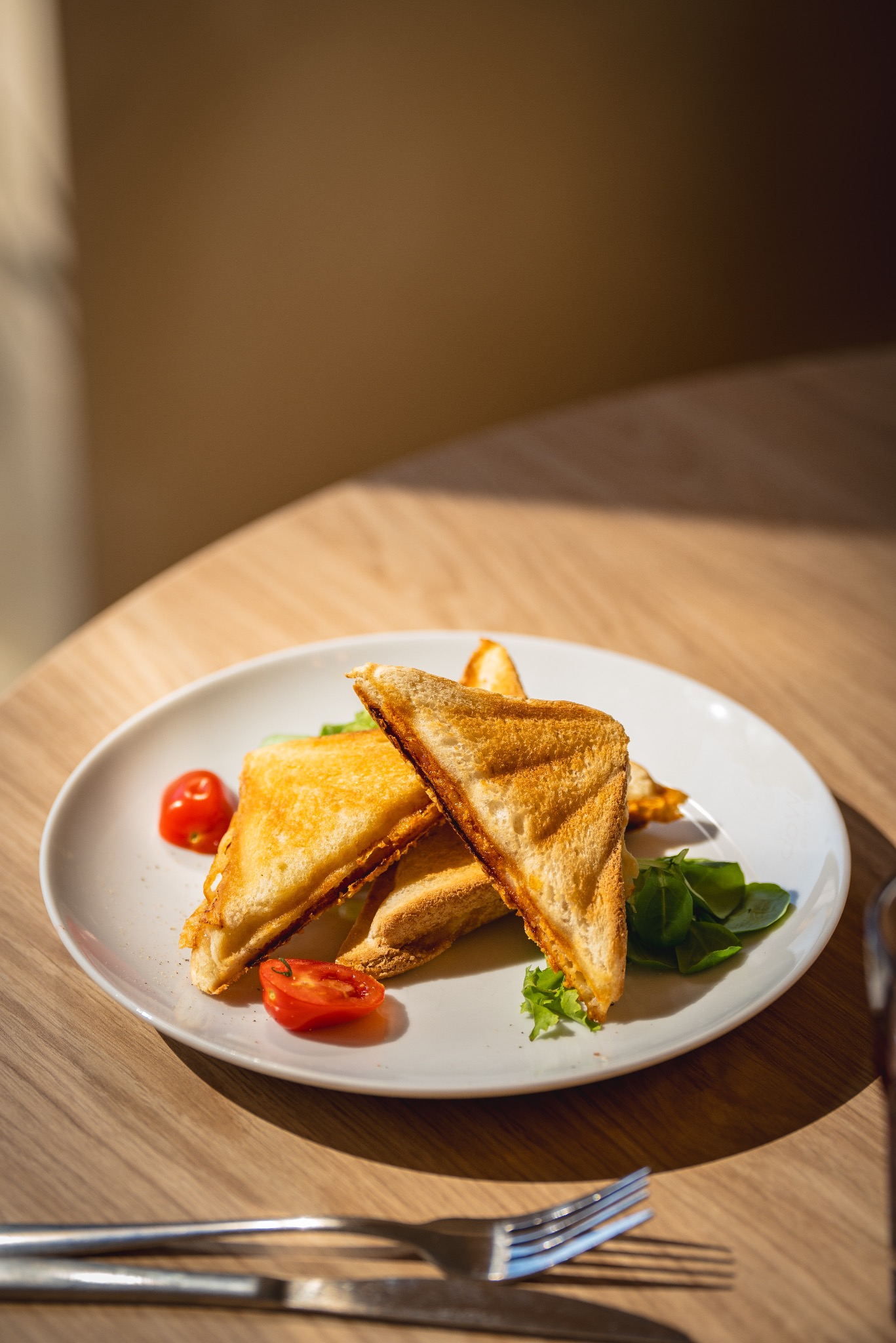 Trójkątne tosty na białym talerzu, udekorowane listkami bazylii i połówkami pomidorków koktajlowych, ułożone na drewnianym stole w promieniach słonecznego światła, z widocznymi sztućcami w dolnej...