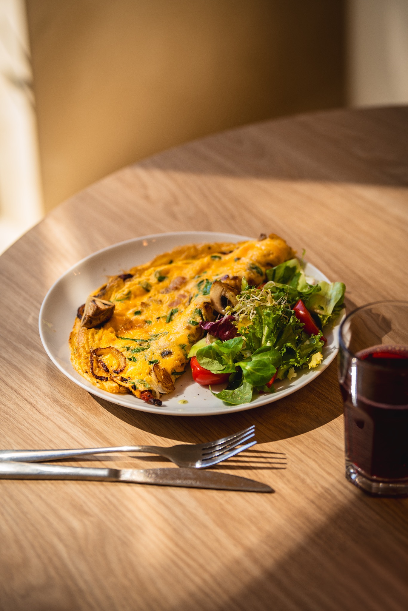 Omeleta z grzybami i szpinakiem podana z sałatką ze świeżych warzyw i szklanką soku na drewnianym stole w ciepłym świetle słonecznym, widelcem i nożem obok.