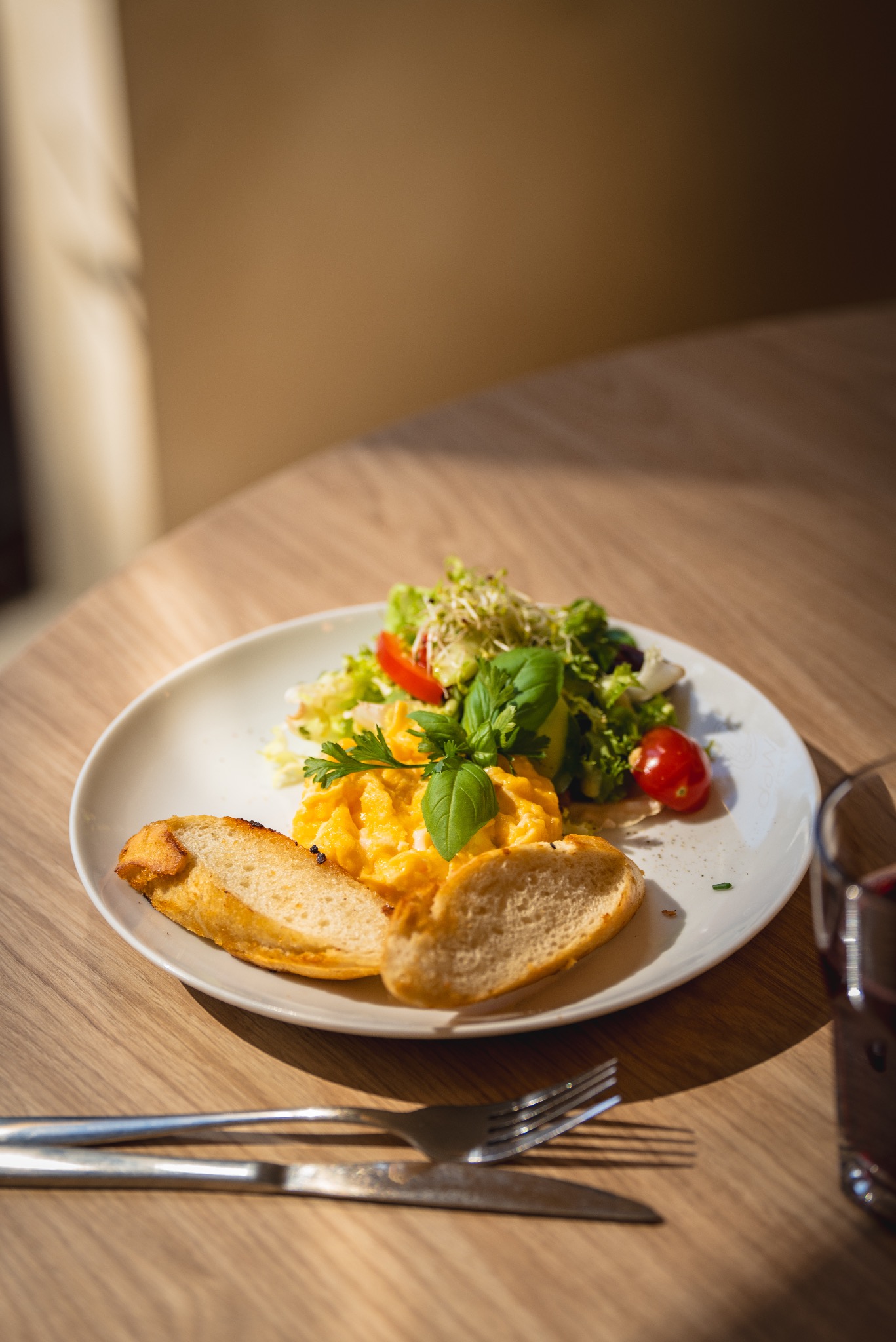 Śniadanie na białym talerzu: jajecznica z ziołami, sałatka ze świeżych warzyw i grzanki, ustawione na drewnianym stole w jasnym świetle słonecznym, obok szklanka z ciemnym napojem oraz sztućce.