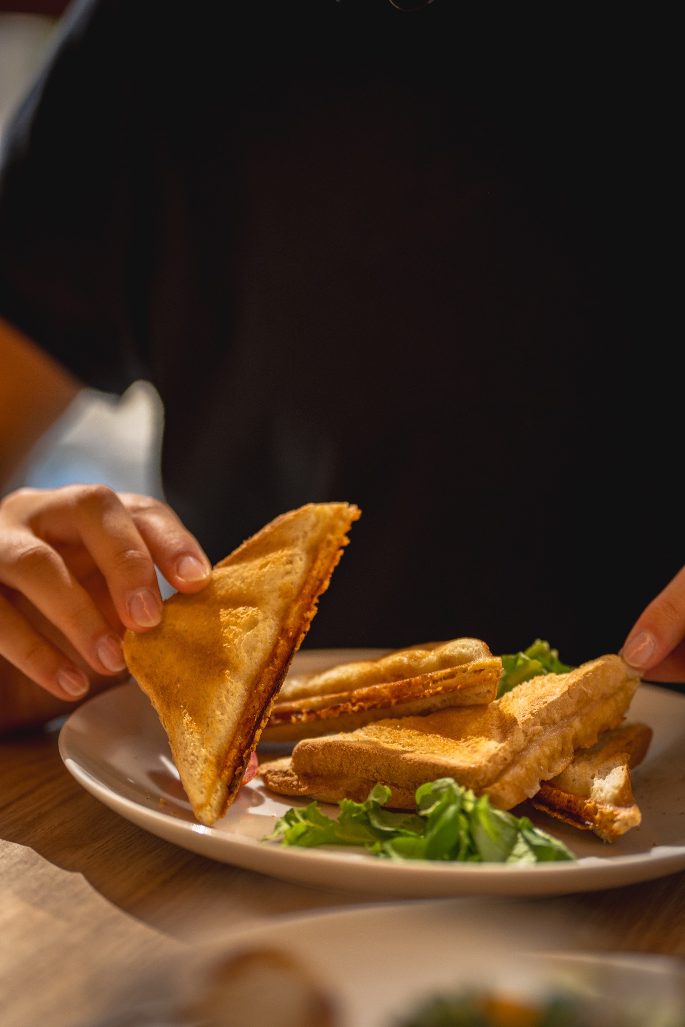 Osoba trzyma trójkątną kanapkę z tostowego chleba z widocznym nadzieniem, obok talerz z innymi kanapkami i zielonymi listkami sałaty.