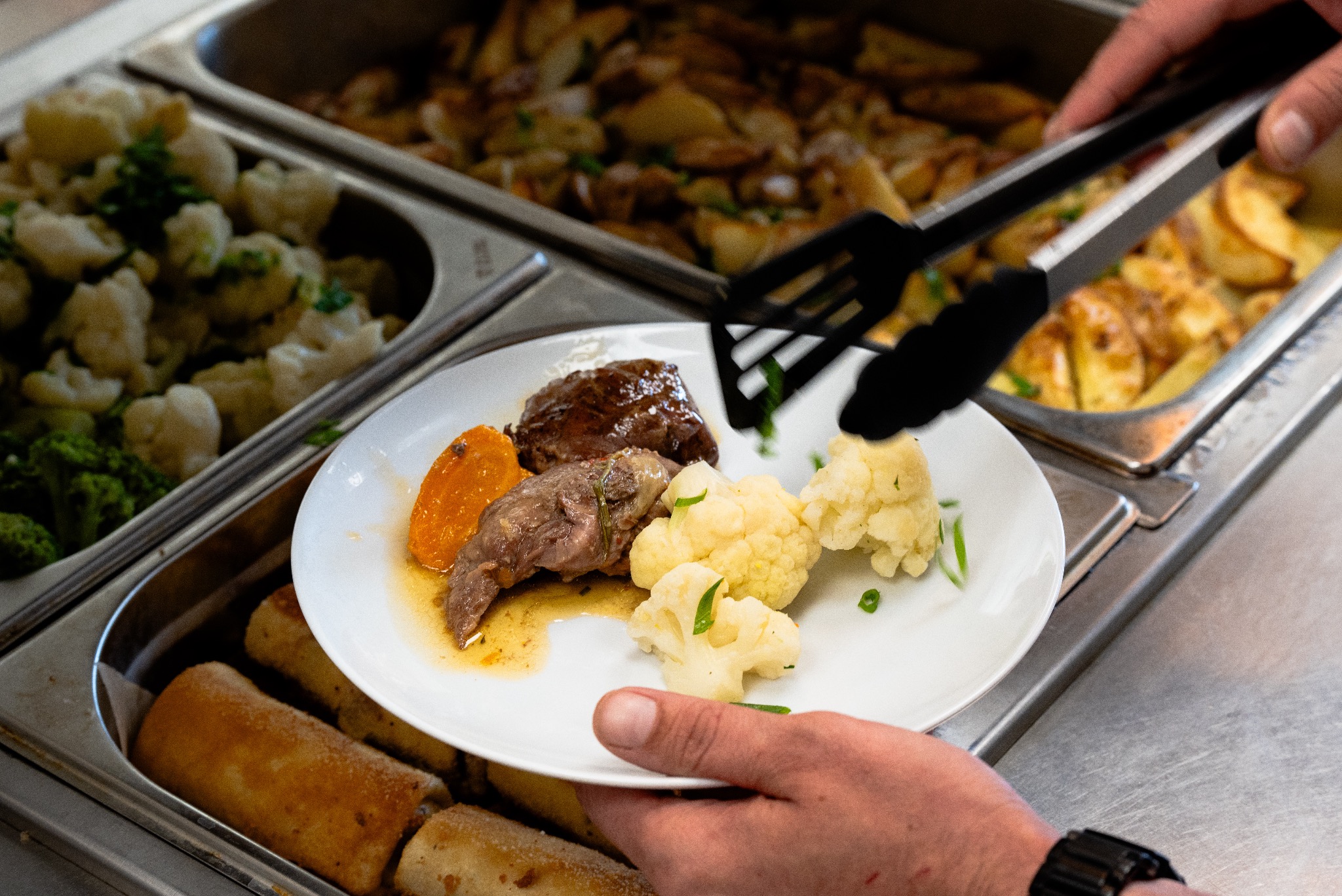 Nakładanie porcji obiadowej na talerz z bemaru: mięso w sosie, gotowany kalafior i pieczona marchewka, widoczne szczypce i inne potrawy w tle.