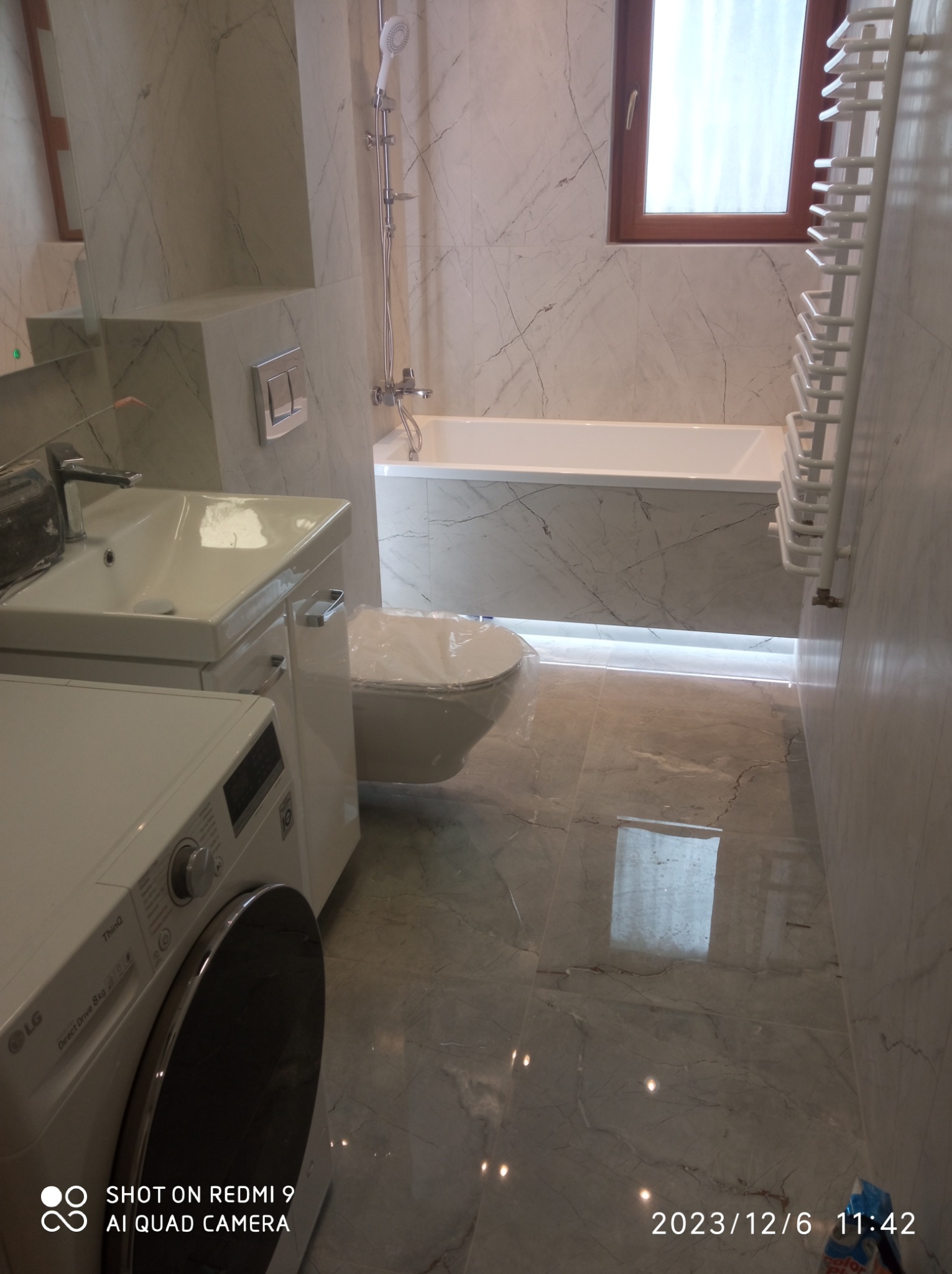 Wykończona łazienka z marmurowymi płytkami, białą armaturą, grzejnikiem drabinkowym i pralką LG z przodu. Umywalka z szafką, podwieszana toaleta i wanna w zabudowie. Widoczne okno z drewnianą ramą.