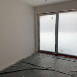 Łukasz Czapiewski - Pokój w trakcie przygotowań do prac wykończeniowych: zabezpieczona folią podłoga, okna osłonięte, surowa ściana, wisząca żarówka. Widok z perspektywy narożnika.