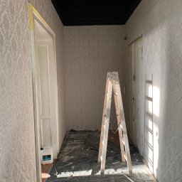 Łukasz Czapiewski - Korytarz po tapetowaniu w jasne wzory. Widoczna drabina, zabezpieczona podłoga i pomalowany na czarno sufit. Taśma malarska zabezpiecza ościeżnice.