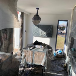 Łukasz Czapiewski - Pokój w trakcie przygotowań do malowania: meble i podłoga zabezpieczone folią malarską, lampa owinięta, widoczne narzędzia i materiały malarskie. Jasne wnętrze z oknem na ogród.