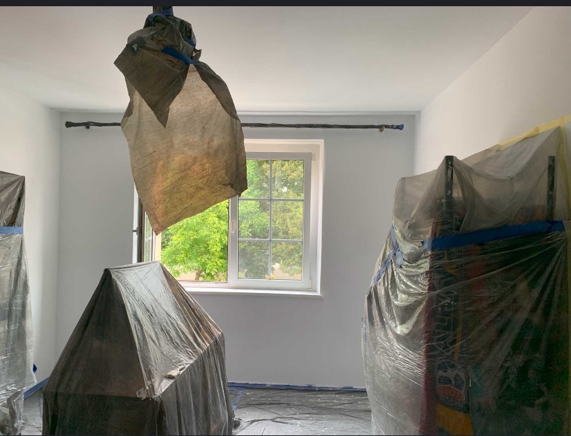 Białe wnętrze pokoju zabezpieczone folią malarską, przygotowane do malowania. Widoczne okno z zielenią na zewnątrz i zakryte meble.