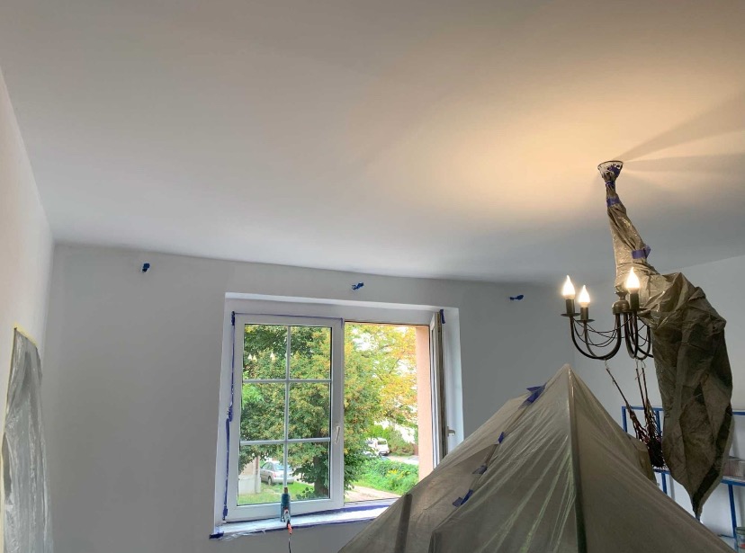 Białe ściany po malowaniu, okno z widokiem na zieleń, zabezpieczony folią żyrandol. Niebieska taśma malarska wokół okna i na ścianach. Świeżo pomalowane wnętrze.