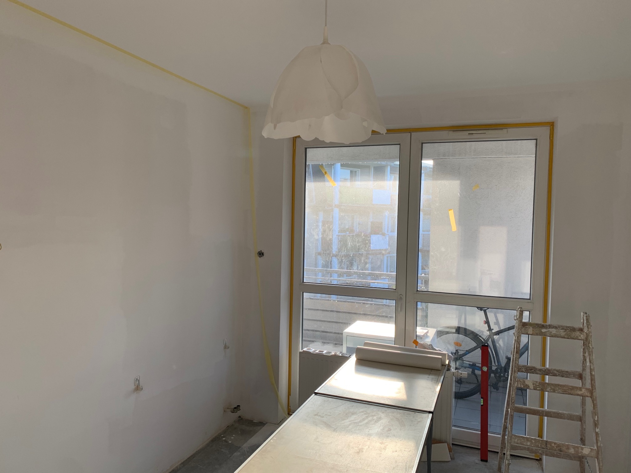 Wnętrze pokoju w trakcie prac wykończeniowych:  ściany przygotowane, zabezpieczone taśmą malarską okna, drabina i stół roboczy.  Widoczny rower za oknem.