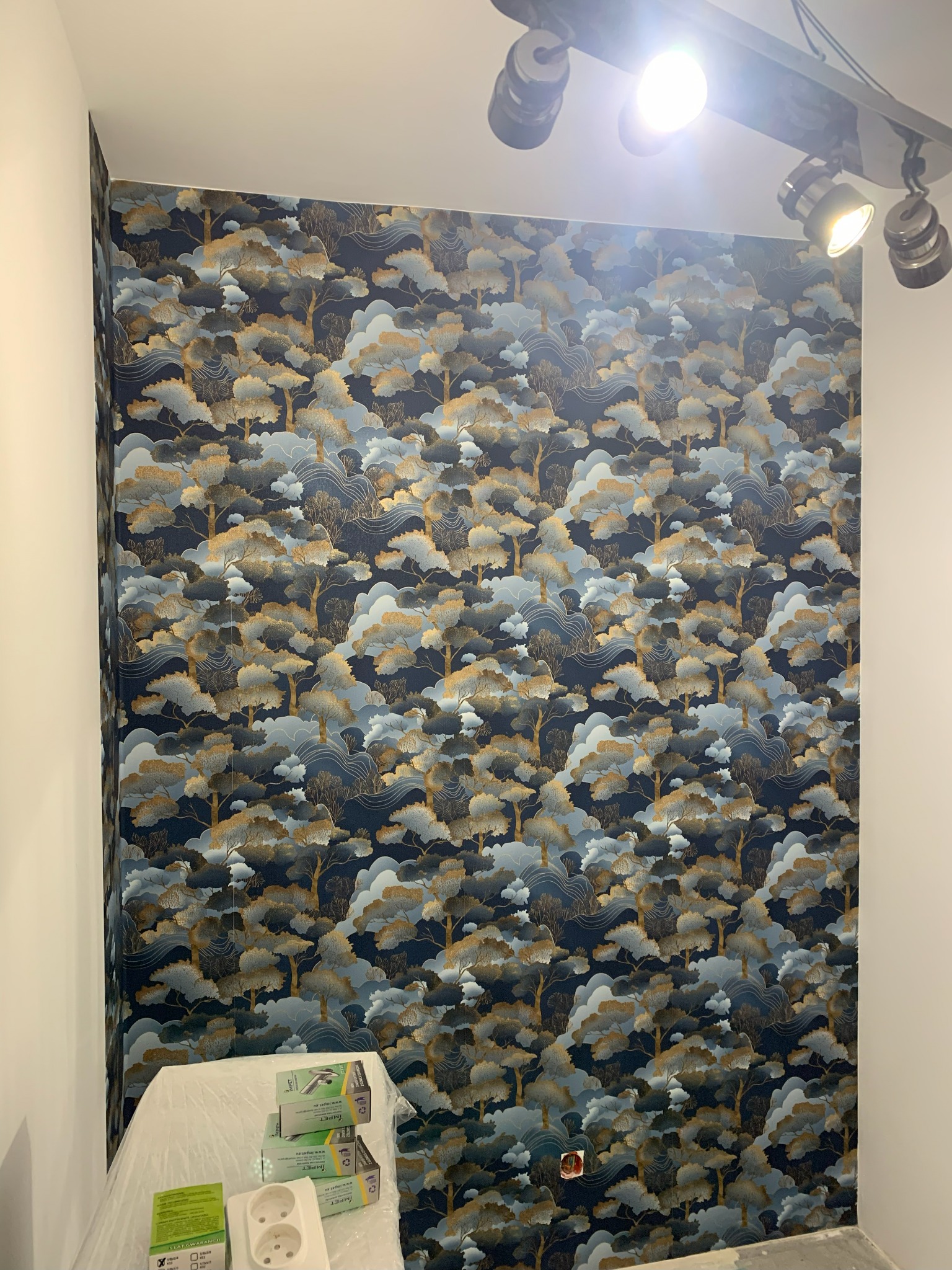 Ściana po tapetowaniu z wzorem drzew na ciemnym tle. Na dole widoczne akcesoria i gniazdko elektryczne. Oświetlenie punktowe na suficie.