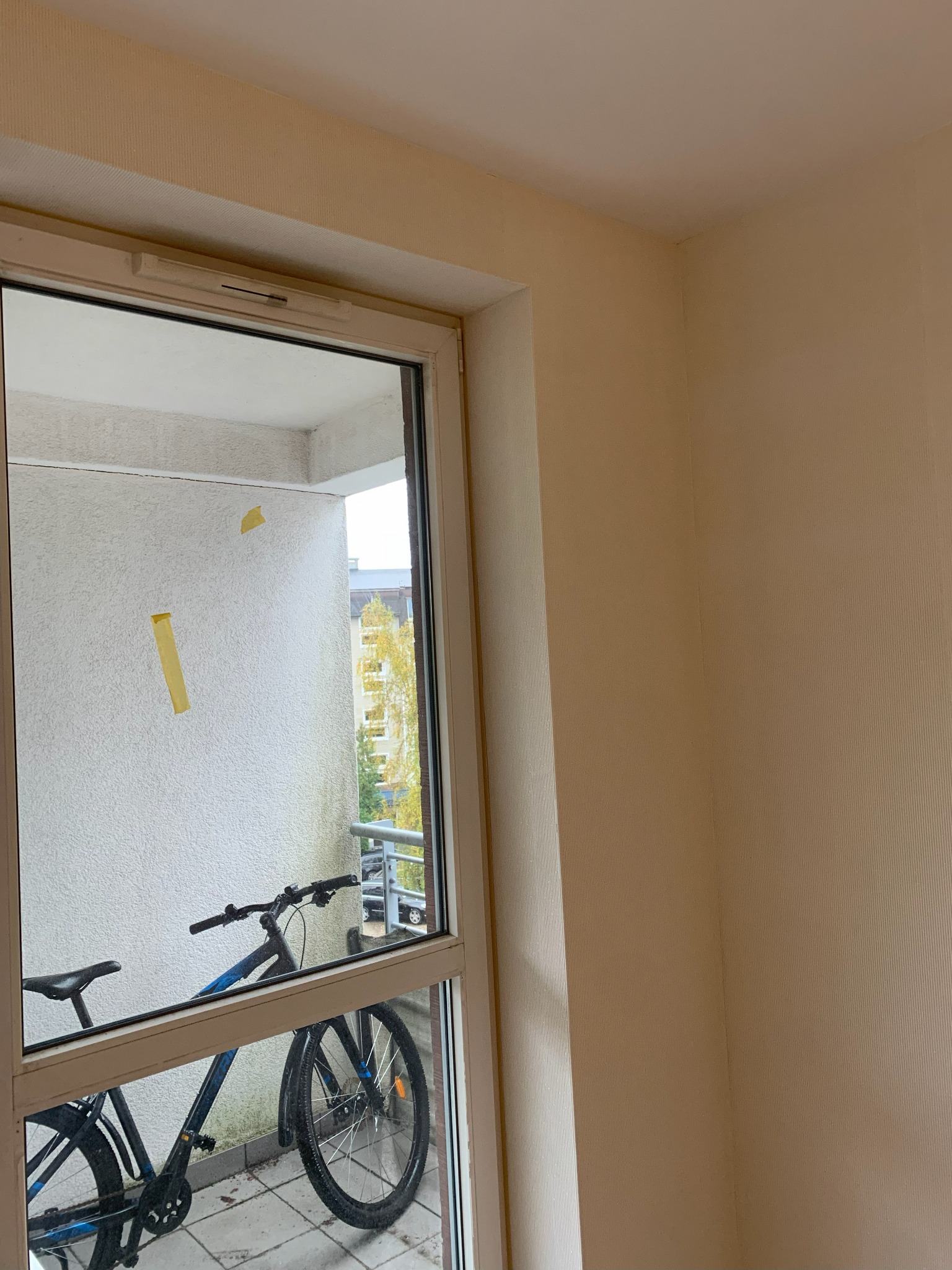 Jasny pokój z widokiem na balkon, gdzie rower oparty jest o ścianę. Na ścianie balkonu widoczne żółte taśmy malarskie.