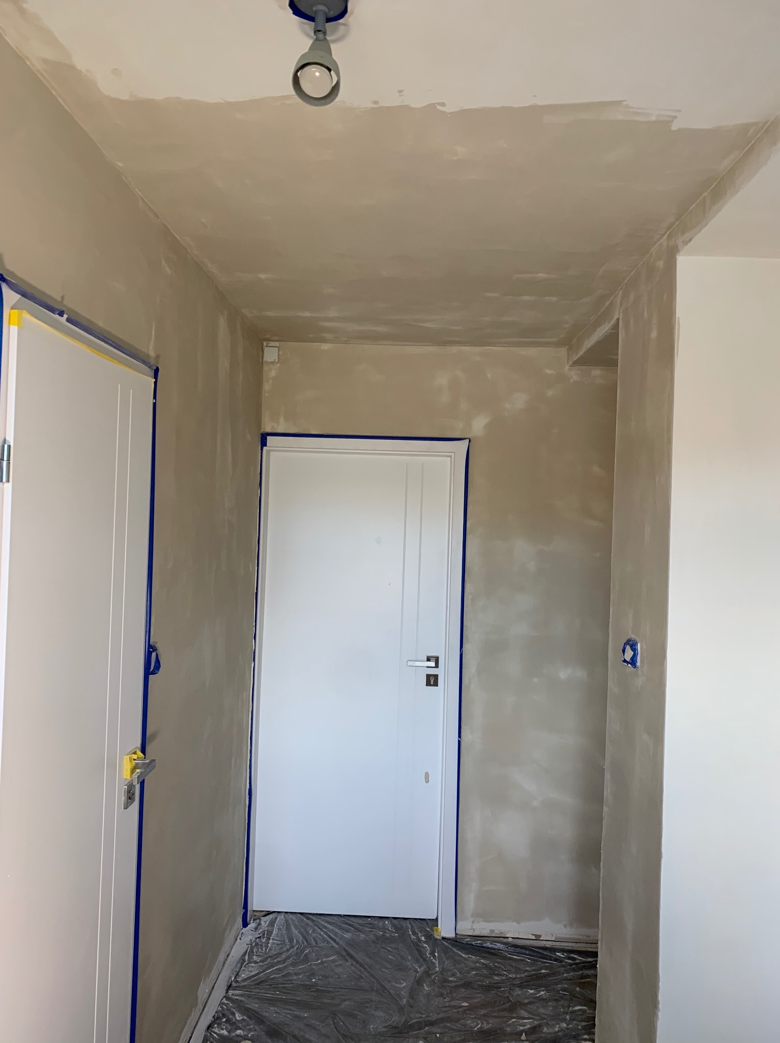 Korytarz w trakcie szpachlowania ścian, zabezpieczone drzwi taśmą malarską, folia ochronna na podłodze, sufit z widocznymi śladami szpachli, lampa sufitowa.