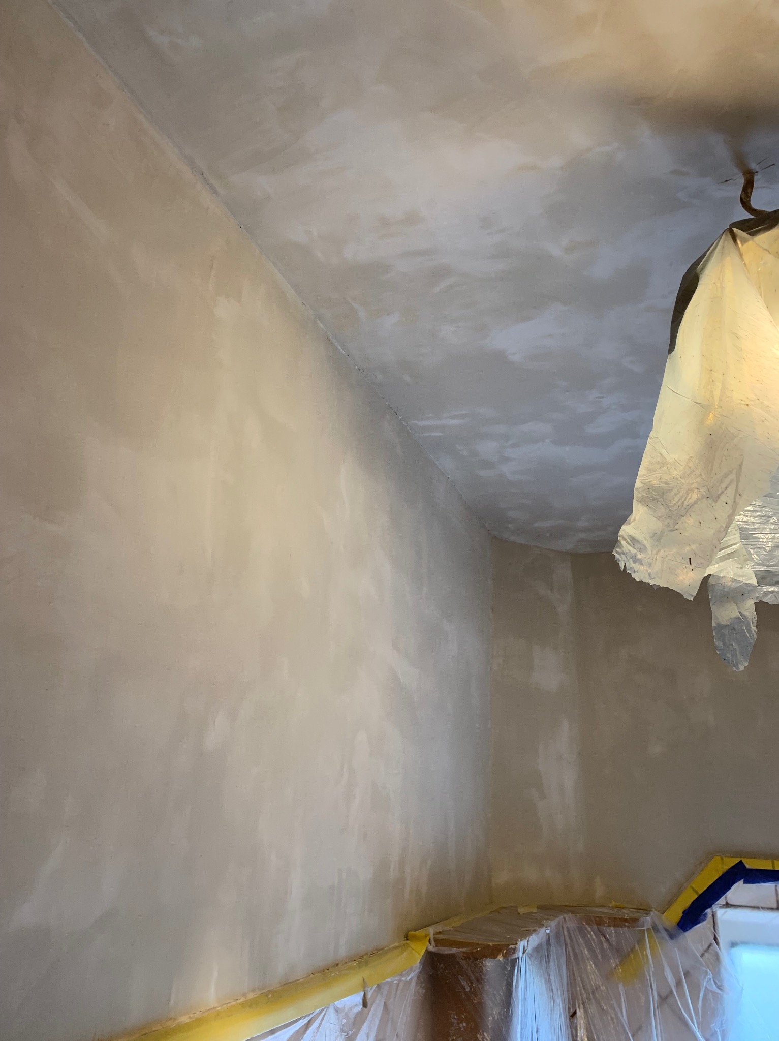 Ściana i sufit po szpachlowaniu, przygotowane do malowania. Zabezpieczone folią i taśmą. Widoczne nierówności gładzi.