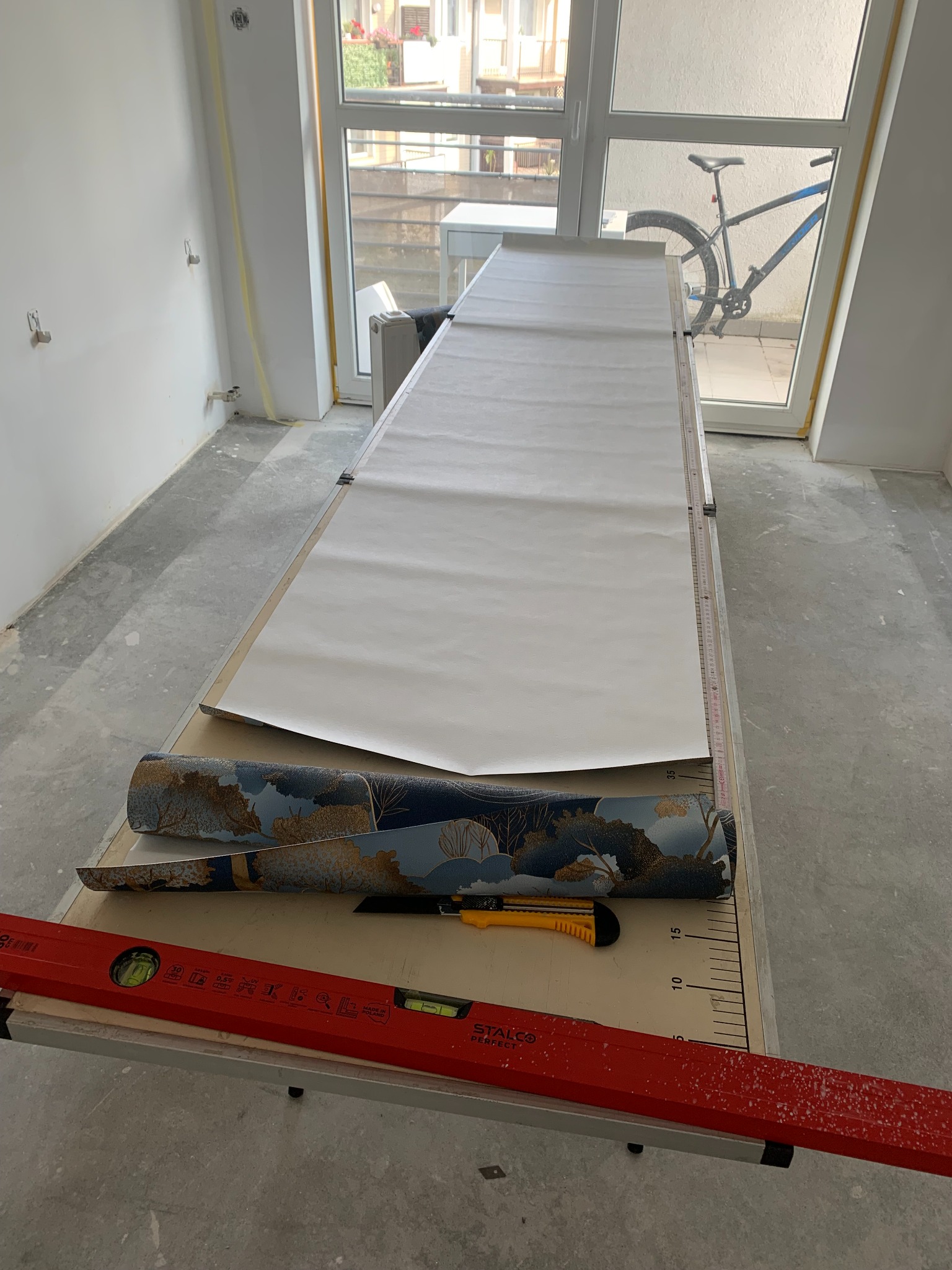 Przygotowanie do tapetowania: rozłożony arkusz tapety na stole z miarką, obok rolka wzorzystej tapety, nóż i poziomica. W tle okno z widocznym rowerem.