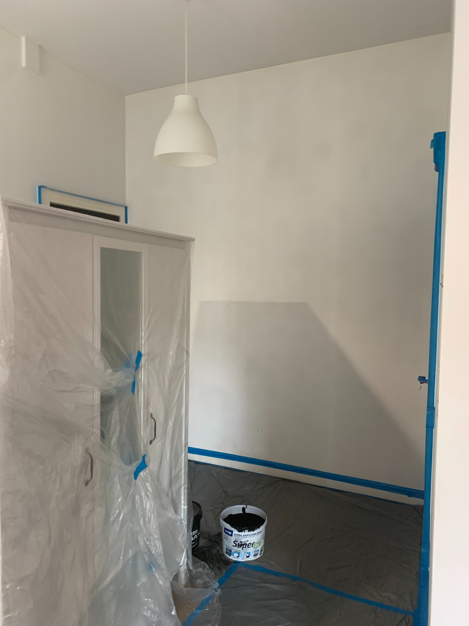 Pokój przygotowany do malowania: zabezpieczone meble folią, ściany i podłoga taśmą malarską, widoczne wiaderka z farbą. Białe ściany, lampa wisząca.