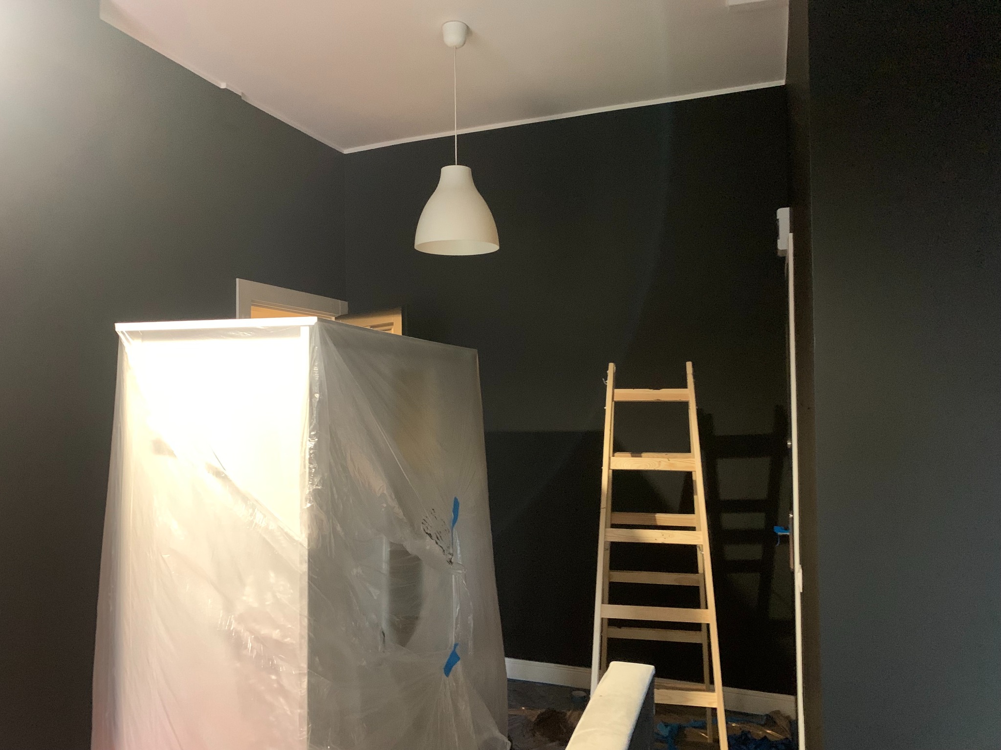 Pokój po malowaniu na ciemny kolor, zabezpieczone meble folią, drabina oparta o ścianę. Widoczna lampa sufitowa. Świeżo pomalowane ściany.