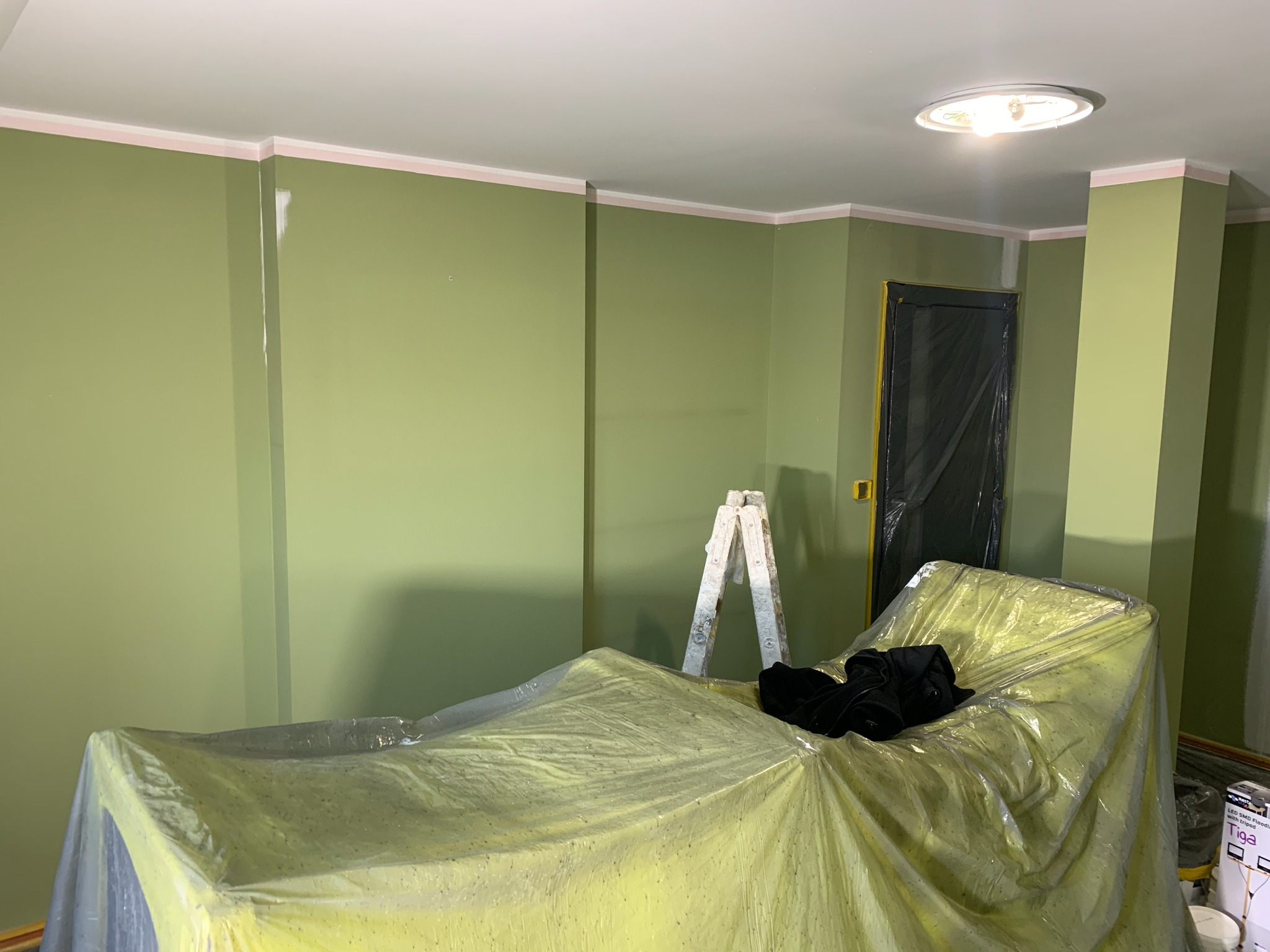 Pokój po malowaniu na zielono, zabezpieczone meble folią, drabina oparta o ścianę, widoczne ślady po szpachlowaniu przy suficie. Oświetlenie sufitowe.