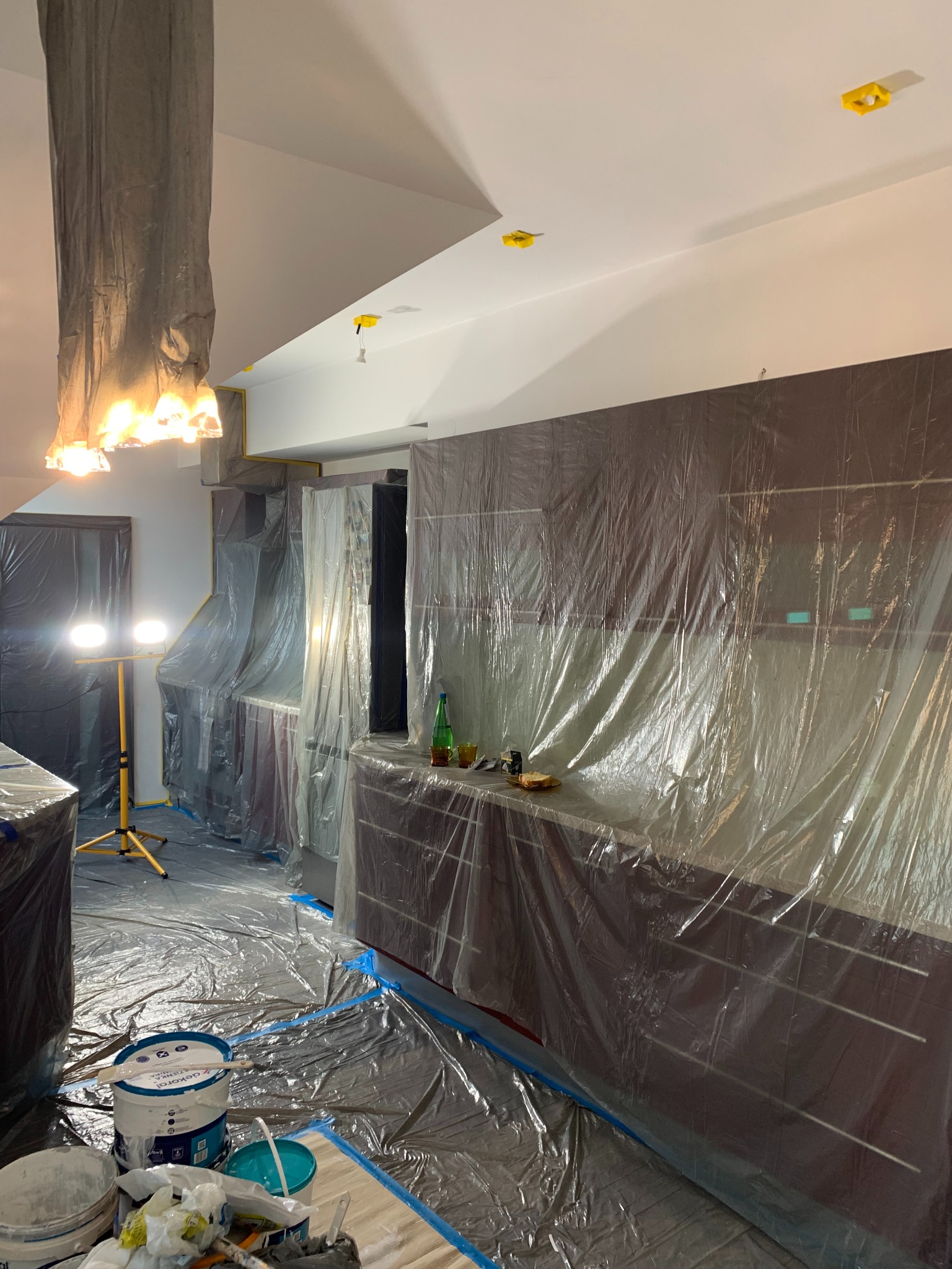 Kuchnia zabezpieczona folią malarską przed pracami wykończeniowymi. Widoczne wiadra z farbą, oświetlenie robocze i zakryte meble. Przygotowanie do malowania.