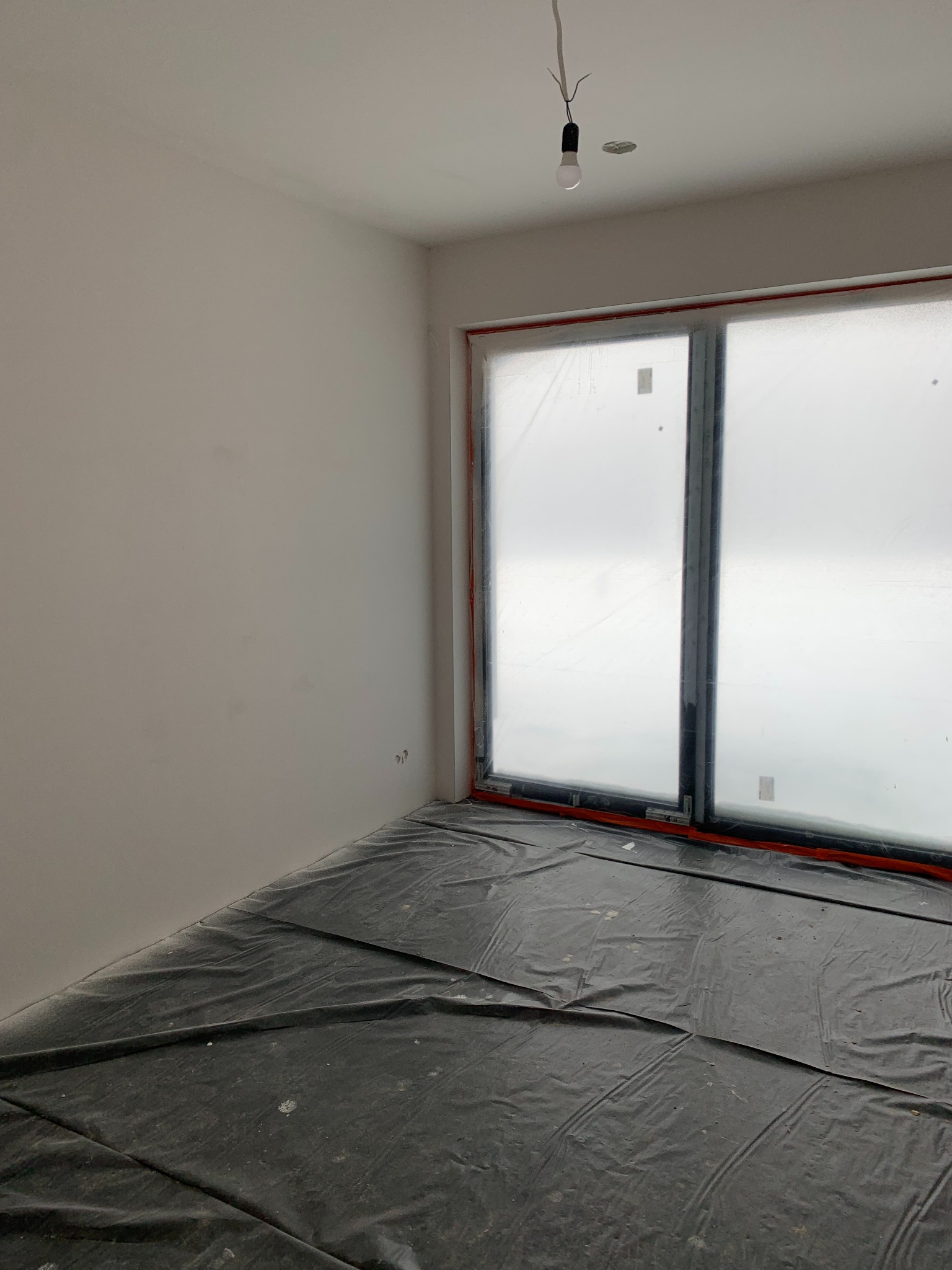 Pokój w trakcie przygotowań do prac wykończeniowych: zabezpieczona folią podłoga, okna osłonięte, surowa ściana, wisząca żarówka. Widok z perspektywy narożnika.
