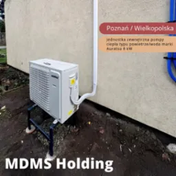 Zewnętrzna jednostka klimatyzacyjna Auratsu 8kW zamontowana na metalowej konstrukcji przy ścianie budynku. Widoczne rury doprowadzające, tło z ziemią i fragmentem roślinności.