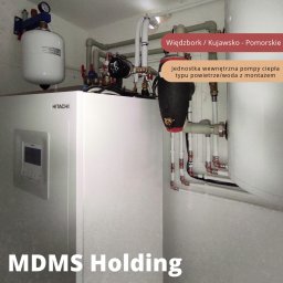 MDMS HOLDING Spółka z o.o. - Wewnętrzna jednostka pompy ciepła Hitachi typu powietrze/woda z orurowaniem i zbiornikiem wyrównawczym, zainstalowana w pomieszczeniu technicznym. Widoczne połączenia hydrauliczne i elementy armatury.