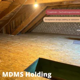 Poddasze z drewnianą konstrukcją dachu, ocieplonym stropem wełną mineralną i wykończonym płytami OSB, widoczny otwór włazowy z zielonym obramowaniem, oświetlone pojedynczą żarówką.