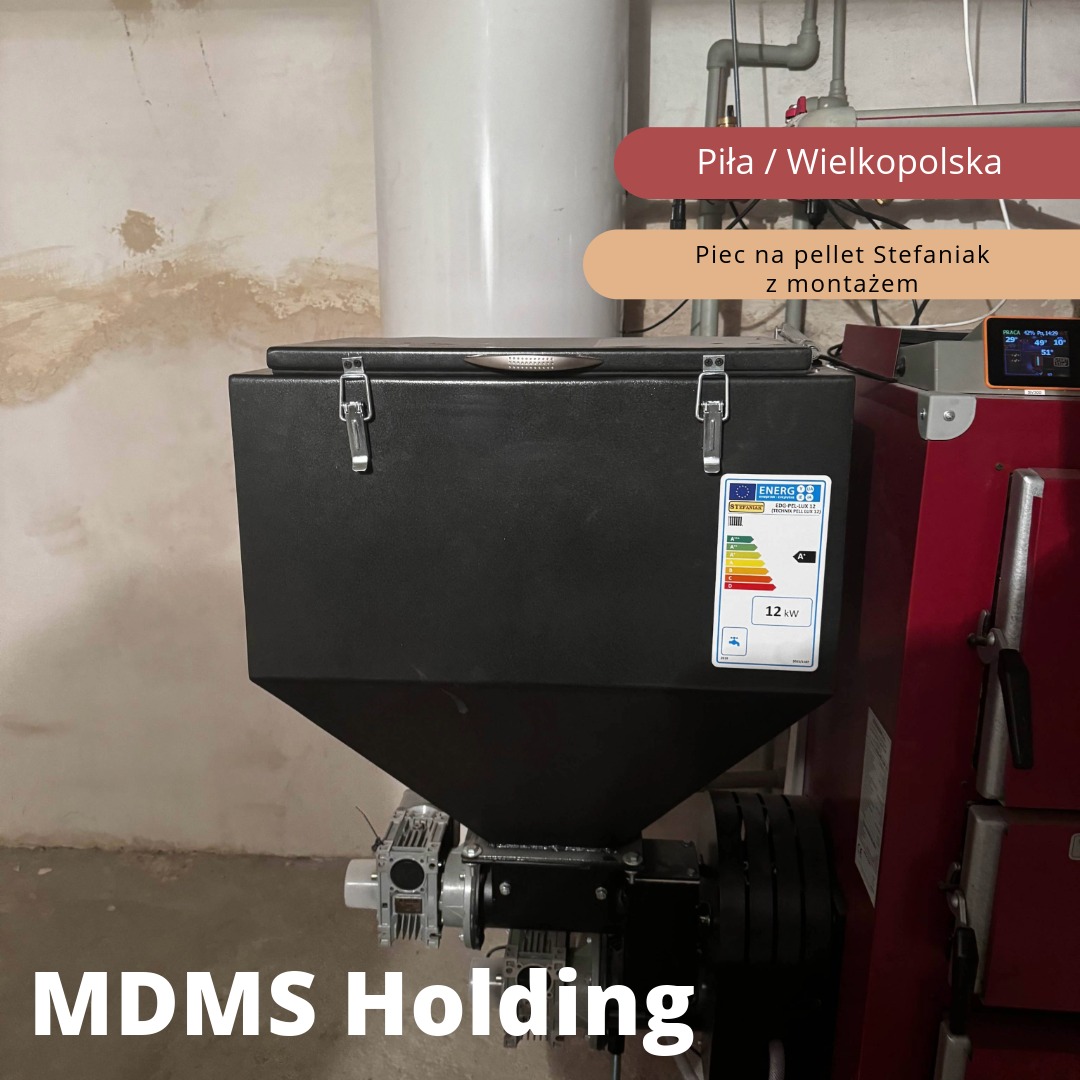 Czarny piec na pellet z podajnikiem, widoczna etykieta energetyczna, zainstalowany obok czerwonego kotła, na tle ściany z widocznymi śladami wilgoci.