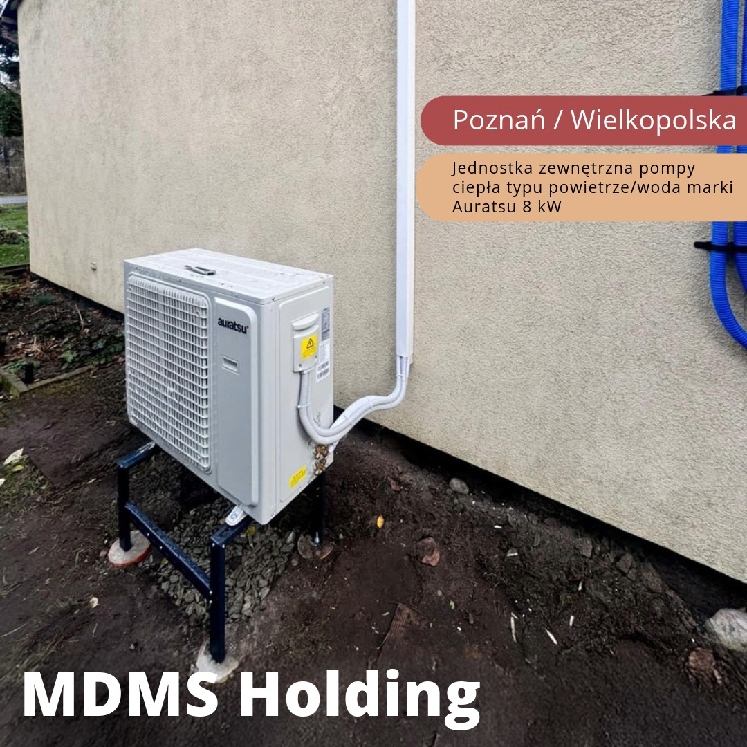Zewnętrzna jednostka klimatyzacyjna Auratsu 8kW zamontowana na metalowej konstrukcji przy ścianie budynku. Widoczne rury doprowadzające, tło z ziemią i fragmentem roślinności.