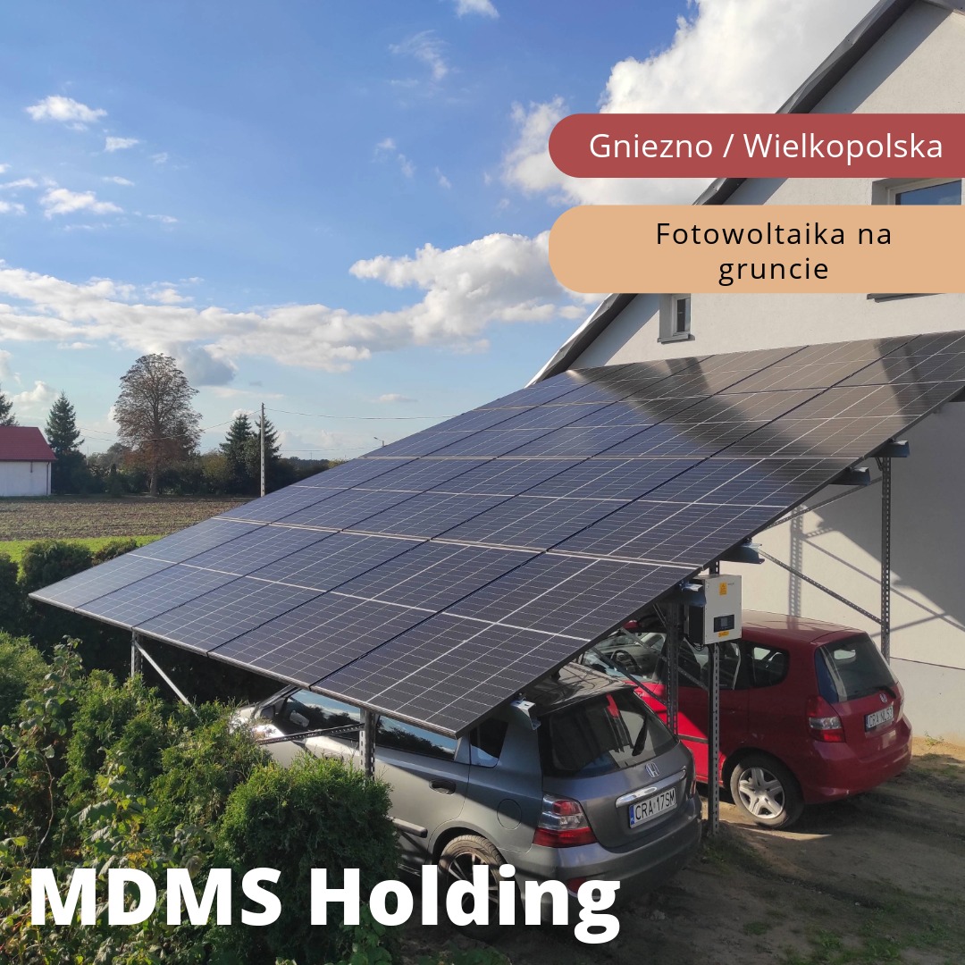 Instalacja paneli słonecznych na gruncie, tworząca zadaszenie dla dwóch samochodów osobowych, widok z boku w słoneczny dzień. Na pierwszym planie krzewy.