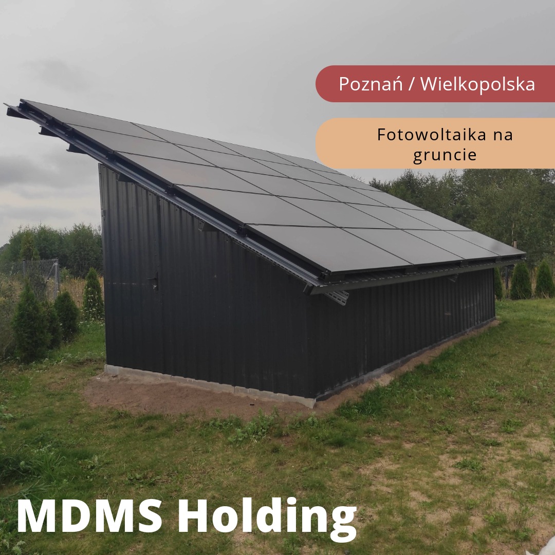 Czarny budynek gospodarczy z panelami słonecznymi na dachu, widok z poziomu gruntu, trawa i drzewa w tle.