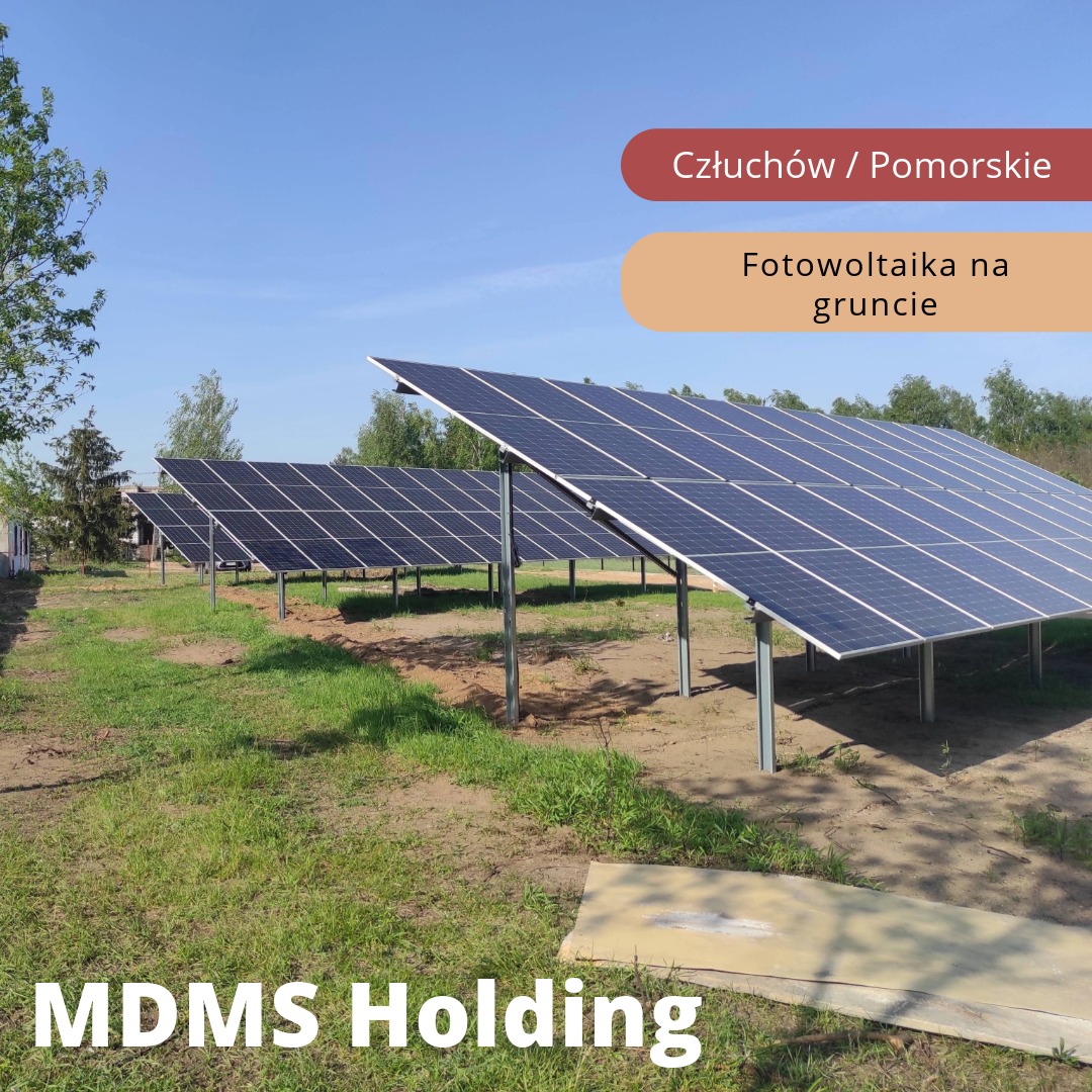 Instalacja paneli słonecznych na gruncie, widok na rzędy paneli w słoneczny dzień, z widocznym trawnikiem i fragmentem drzewa po lewej stronie kadru. Na pierwszym planie betonowa płyta.