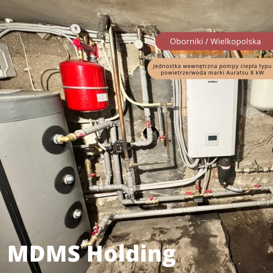 Wewnętrzna instalacja grzewcza z zbiornikiem akumulacyjnym i białą jednostką wewnętrzną 8kW marki Auratsu, liczne rury i zawory, ściany piwniczne w tle.