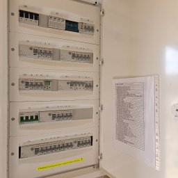 Mateusz Mrugała - Otwarta skrzynka rozdzielcza z wyłącznikami i listą obwodów, widoczne zabezpieczenia Eaton, opis obwodów w koszulce. Żółta naklejka ostrzegawcza.