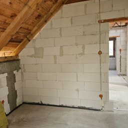 Mateusz Mrugała - Surowy stan poddasza z drewnianą więźbą, ściany z betonu komórkowego, widoczne przewody elektryczne i przygotowane otwory pod gniazdka. W tle widoczne kolejne pomieszczenia.