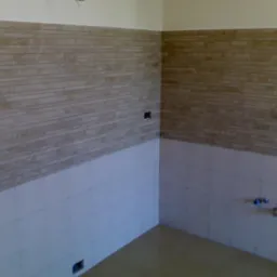 Wykończona ściana z dwoma rodzajami płytek: beżowe poziome paski na górze i białe kwadraty na dole. Widoczne gniazdka elektryczne i rury wodne.