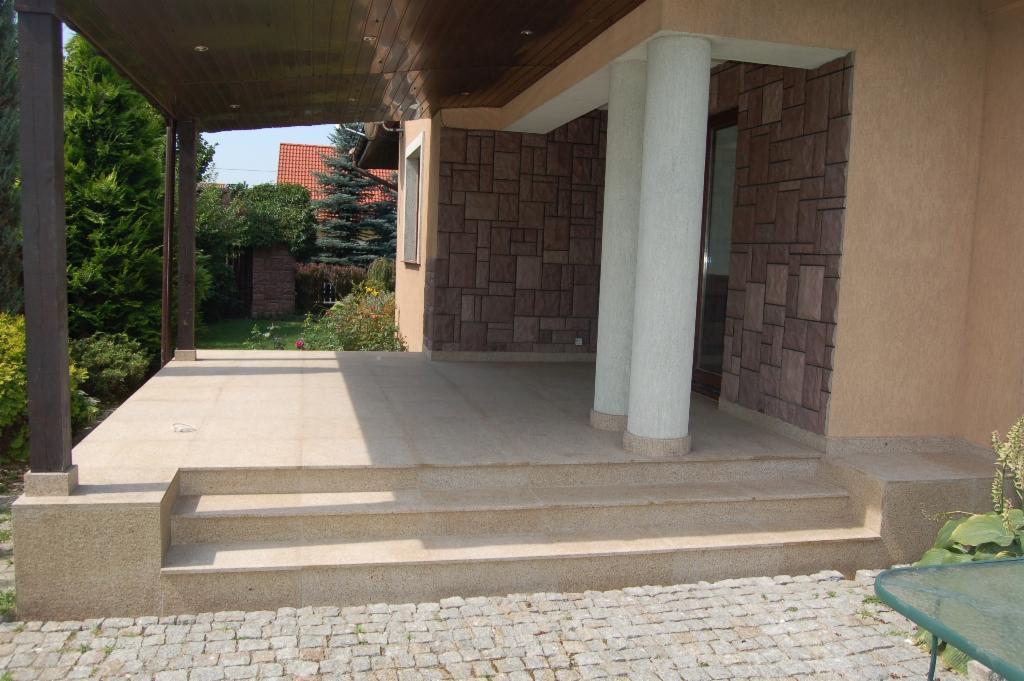 Wykończony taras z kamienną okładziną ściany i kolumnami, widok na ogród. Ułożona kostka brukowa na podjeździe. Drewniane zadaszenie tarasu.