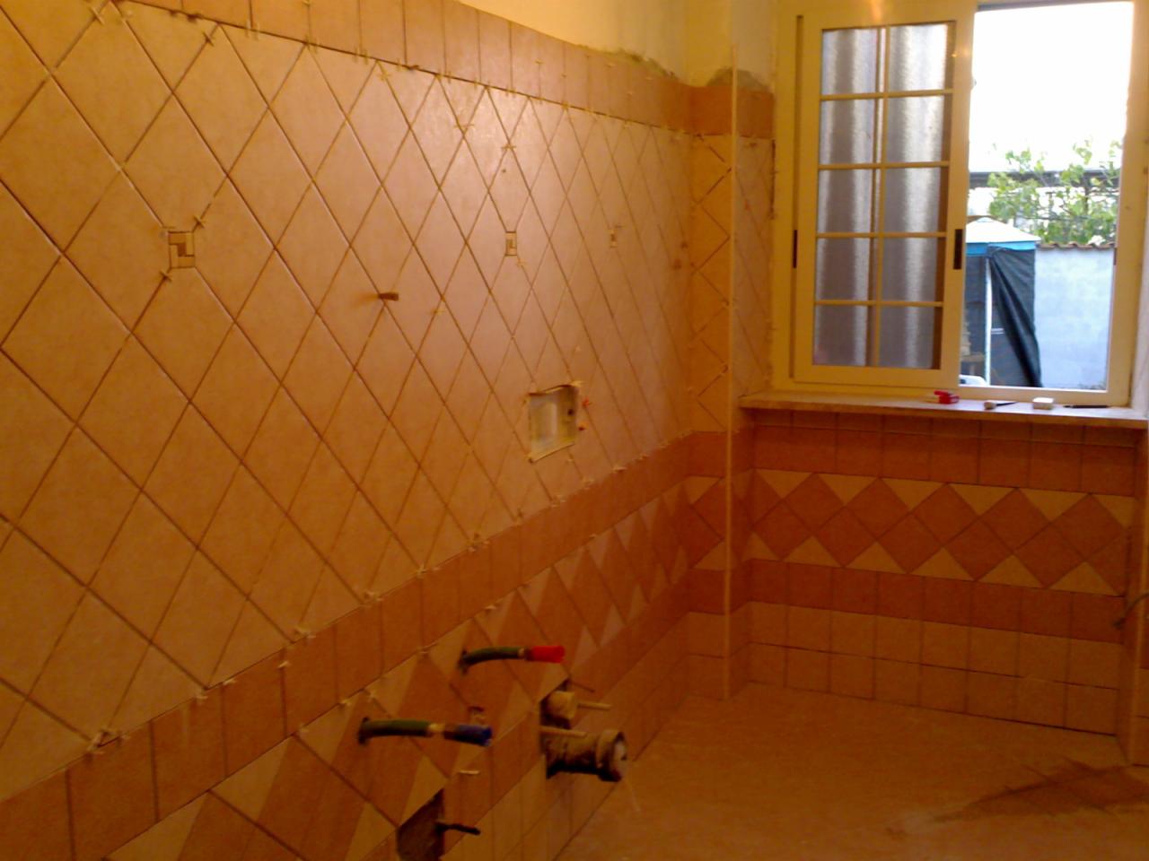 Układanie beżowych płytek w romby na ścianie łazienki, widoczne wystające rury i okno z widokiem na zewnątrz. Częściowo zakończone prace.