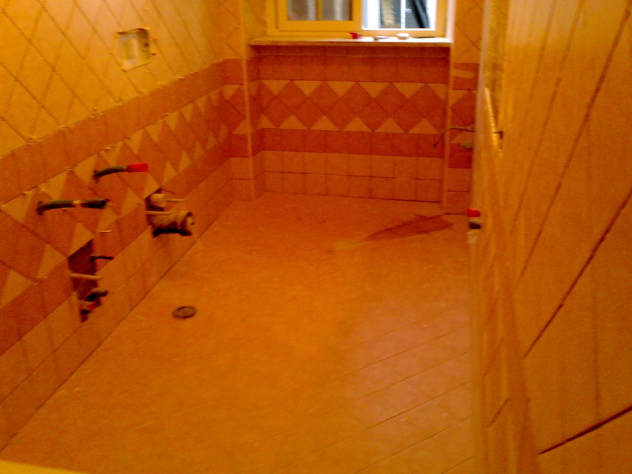 Wykończona łazienka z ułożonymi, pomarańczowymi płytkami na ścianach i podłodze. Widoczne wystające rury instalacji wodnej. Okno z białymi ramami w tle.