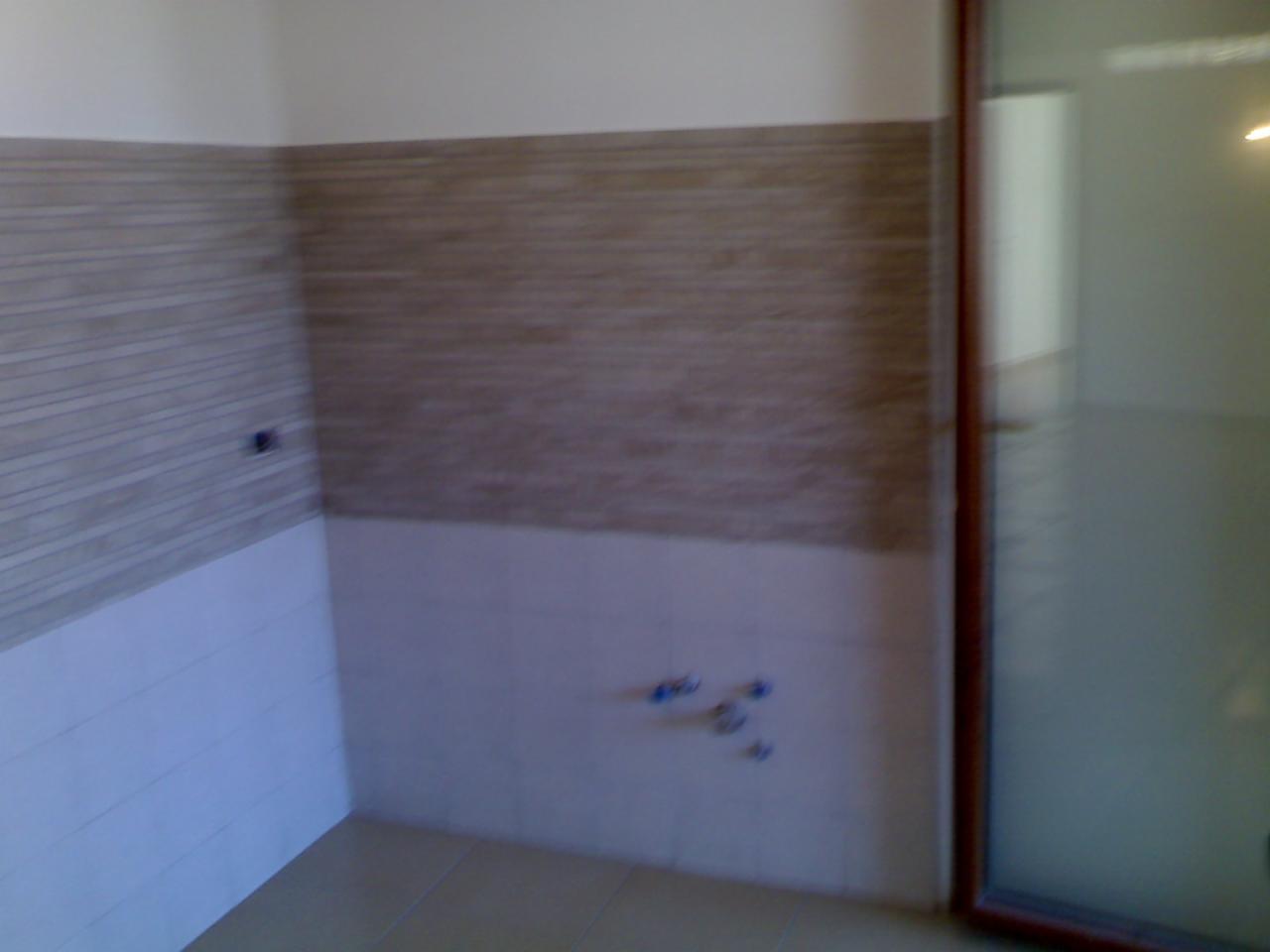 Wykończona ściana w łazience z ułożonymi płytkami w dwóch odcieniach beżu, widoczne przyłącza wodne. Podłoga wyłożona płytkami w jasnym odcieniu. Częściowo widoczne drzwi.