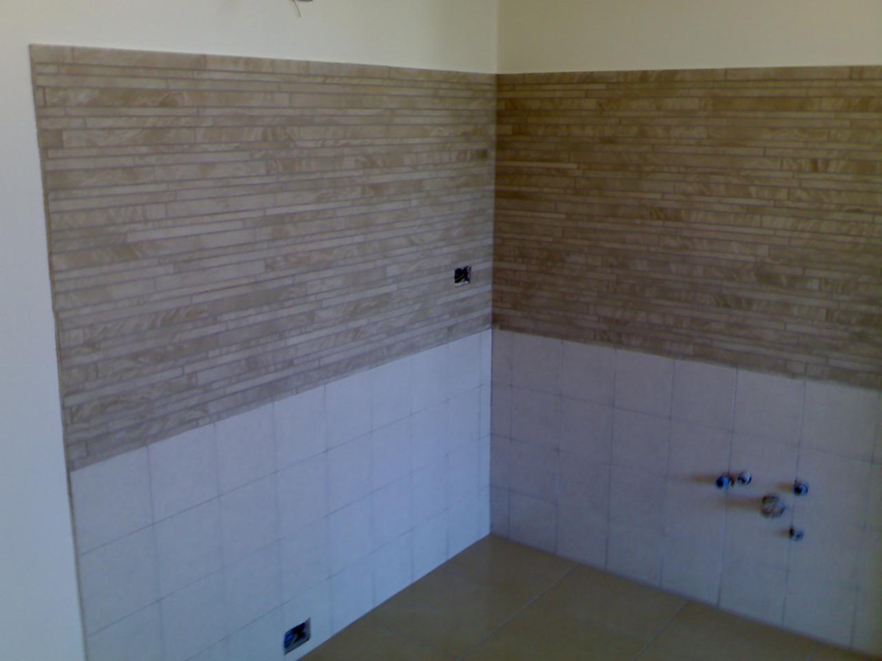 Wykończona ściana z dwoma rodzajami płytek: beżowe poziome paski na górze i białe kwadraty na dole. Widoczne gniazdka elektryczne i rury wodne.