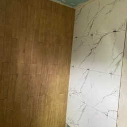 Mateusz Roll - Wnętrze łazienki w trakcie wykańczania: na jednej ścianie ułożone panele imitujące drewno, na drugiej białe płytki z szarym marmurkowym wzorem, widoczne krzyżyki dystansowe, sufit przygotowany...