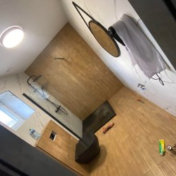 Mateusz Roll - Nowoczesna łazienka po remoncie z drewnopodobną podłogą i ścianą, kabiną prysznicową typu walk-in z czarnymi elementami, okrągłym lustrem w czarnej ramie, wiszącą toaletą i widocznymi narzędziami...