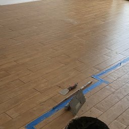 Mateusz Roll - Układanie podłogi z płytek imitujących drewno, widoczne narzędzia: paca z zaprawą, szpachelka, poziomnica i wiadro z klejem, zabezpieczone taśmą malarską.