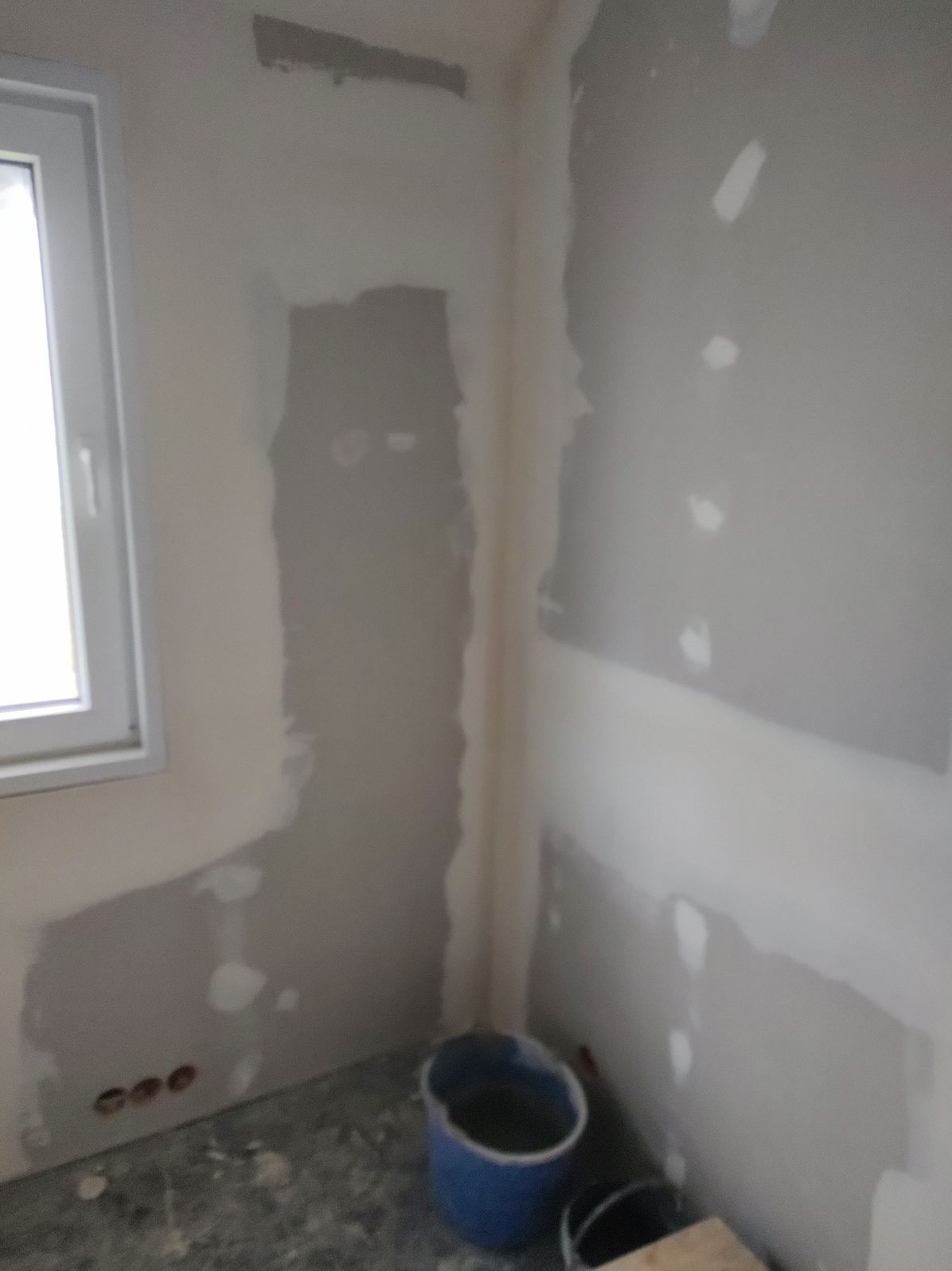 Wnętrze w trakcie prac wykończeniowych: ściany z płyt kartonowo-gipsowych, częściowo zaszpachlowane, widoczne puszki elektryczne, wiadro z zaprawą i narzędzia.