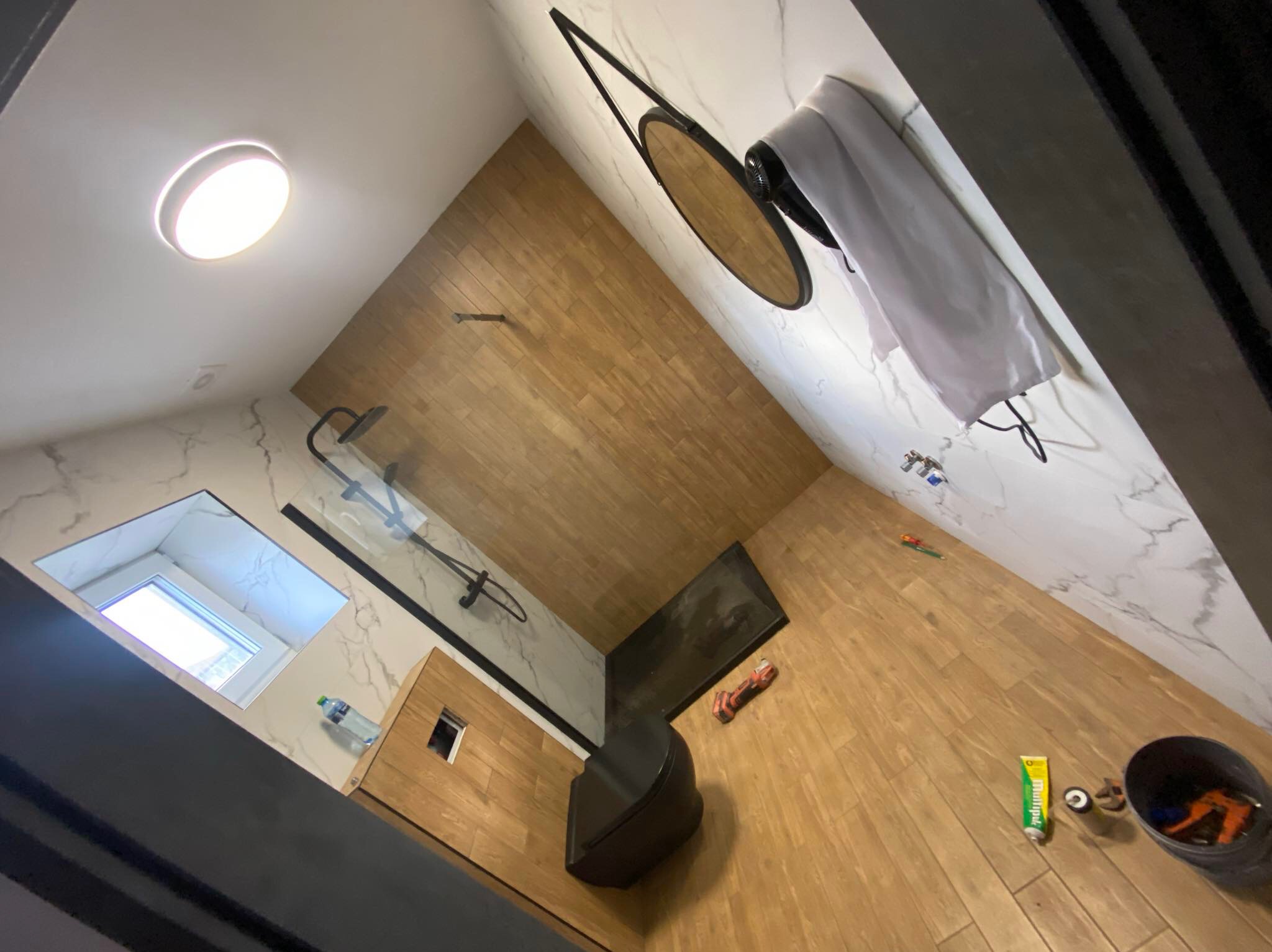 Nowoczesna łazienka po remoncie z drewnopodobną podłogą i ścianą, kabiną prysznicową typu walk-in z czarnymi elementami, okrągłym lustrem w czarnej ramie, wiszącą toaletą i widocznymi narzędziami...